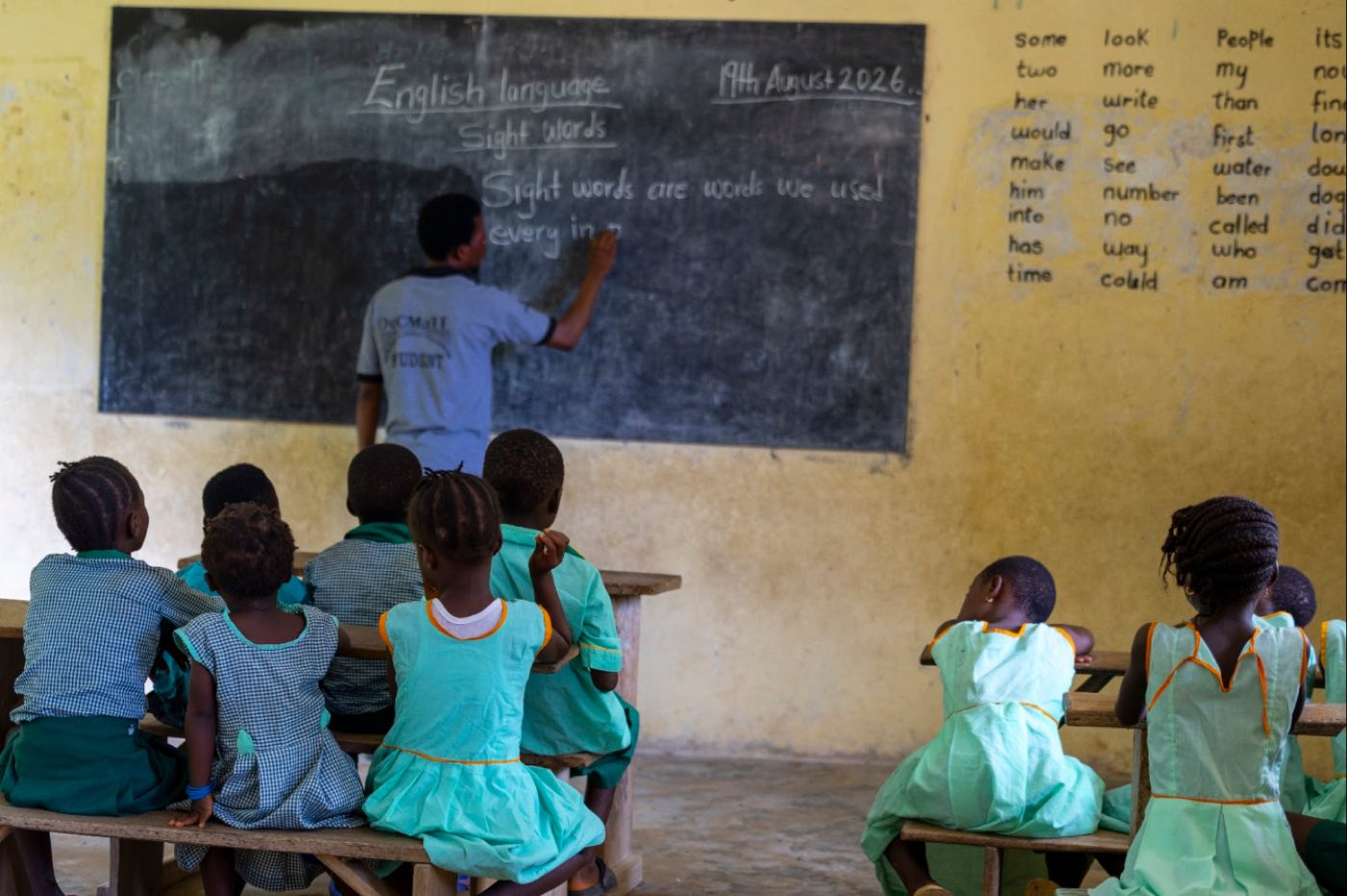 Teacher writing an English lesson on the blackboard, pupils seen from behind